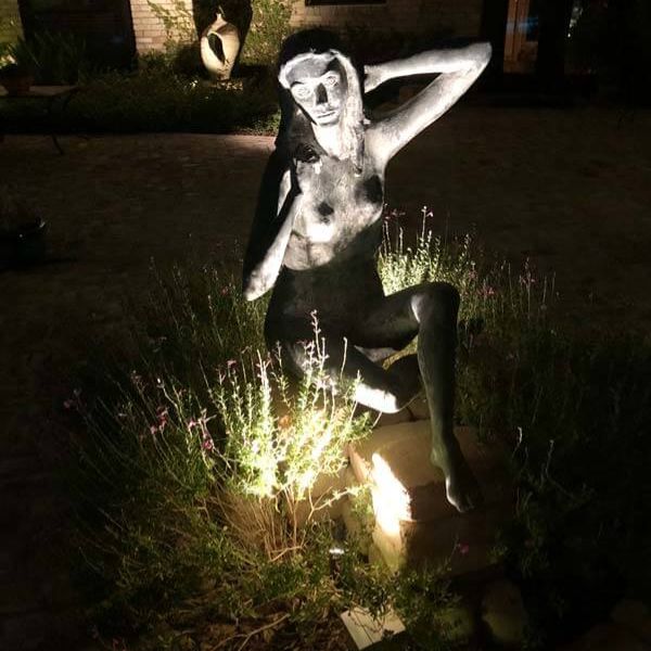 Seated sculpture in outdoor garden at night, lit from below and surrounded by flowers and plants