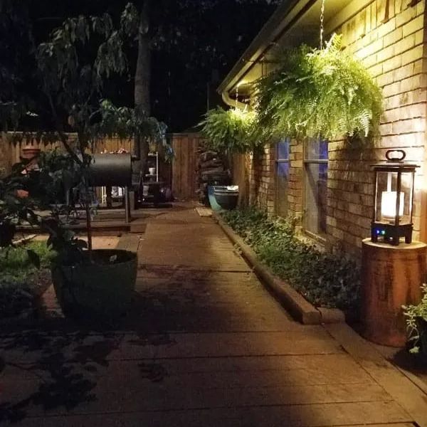 Nighttime patio with brick wall, hanging ferns, lantern on stand, and paved walkway lined with potted plants